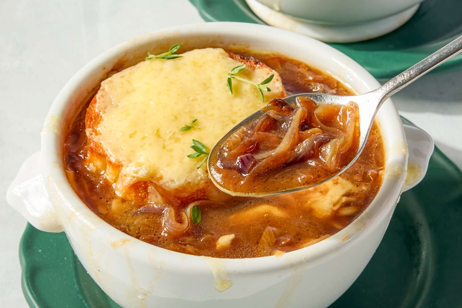 A bowl of French onion soup topped with a slice of melted cheese and garnished with herbs served with a spoon on a green plate