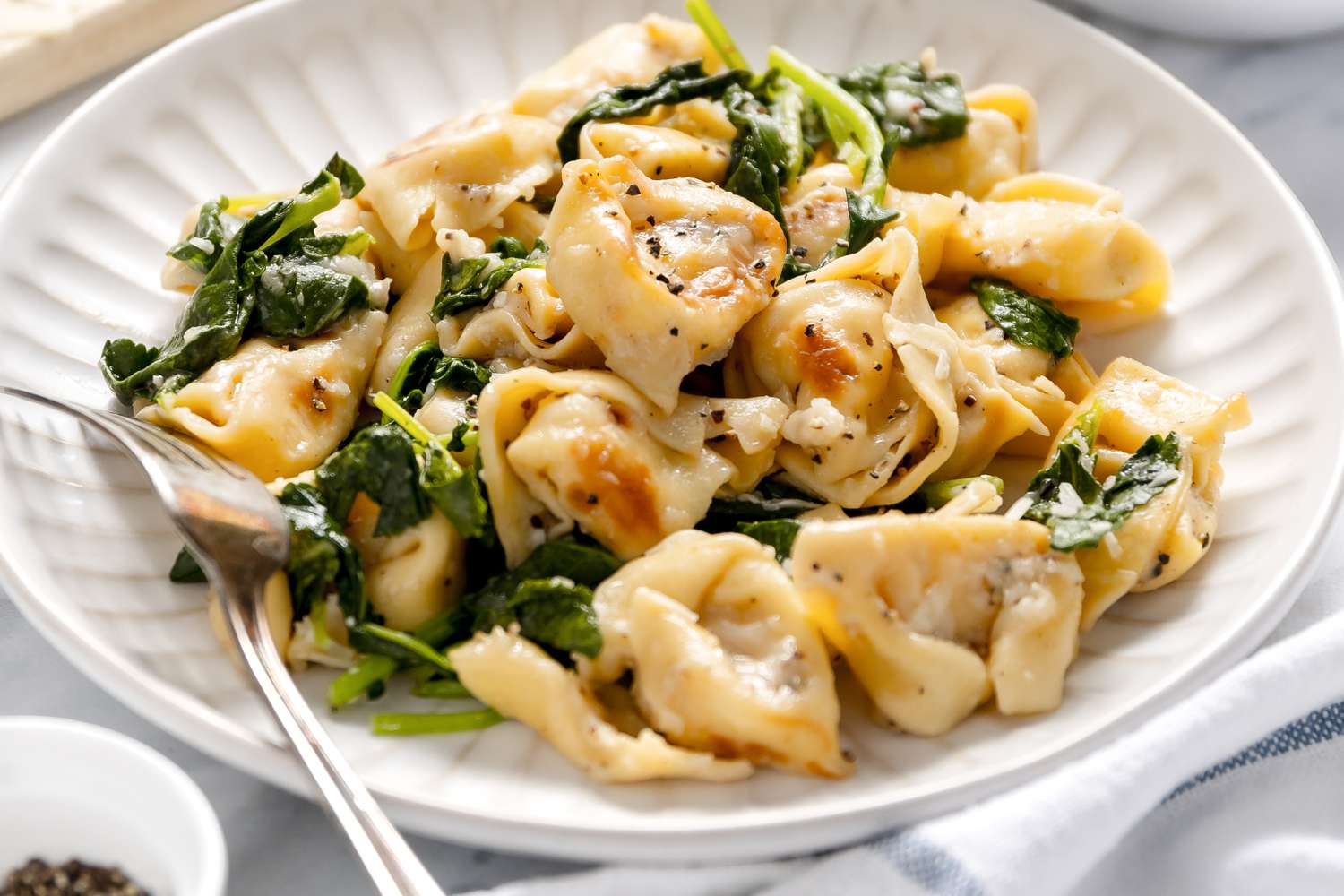 Skillet Cacio e Pepe Tortellini With Wilted Greens on a Plate With a Fork, and in the Surroundings, a Bowl of Cracked Pepper, a Kitchen Towel, and a Cheese Board