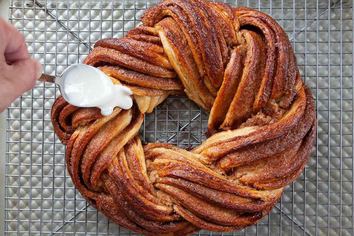 A spoon is drizzling icing on a homemade, golden brown King Cake resting on a cooling rack.