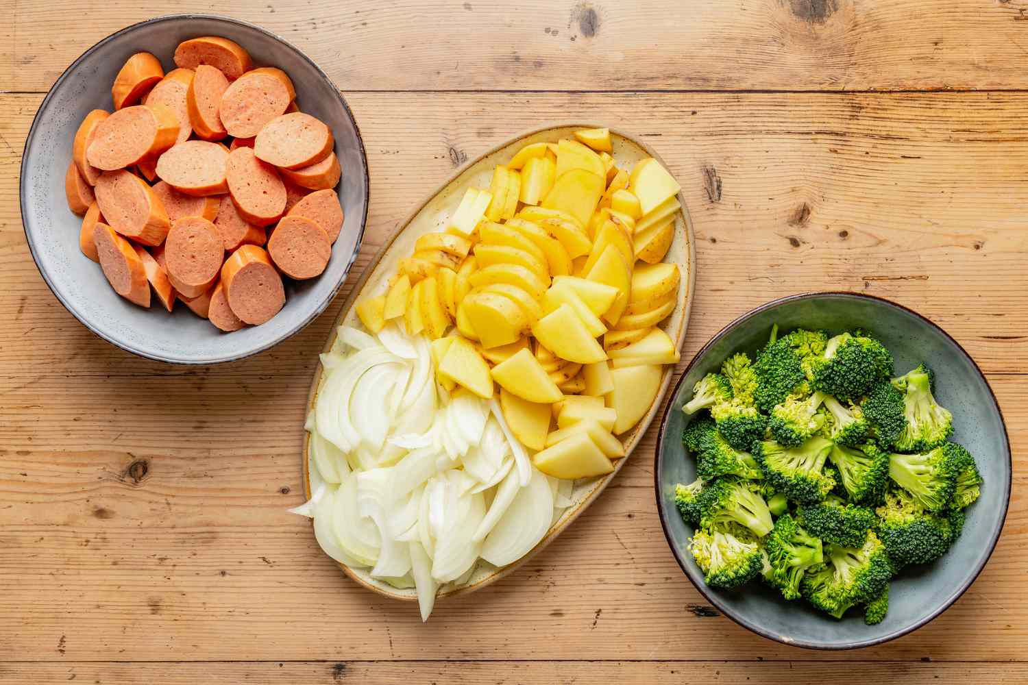 Overhead view of a gray bowl of sliced kielbasa, a bowl of broccoli florets and a brown plate of onion and potato slices for Kielbasa, Potato, and Broccoli Skillet recipe