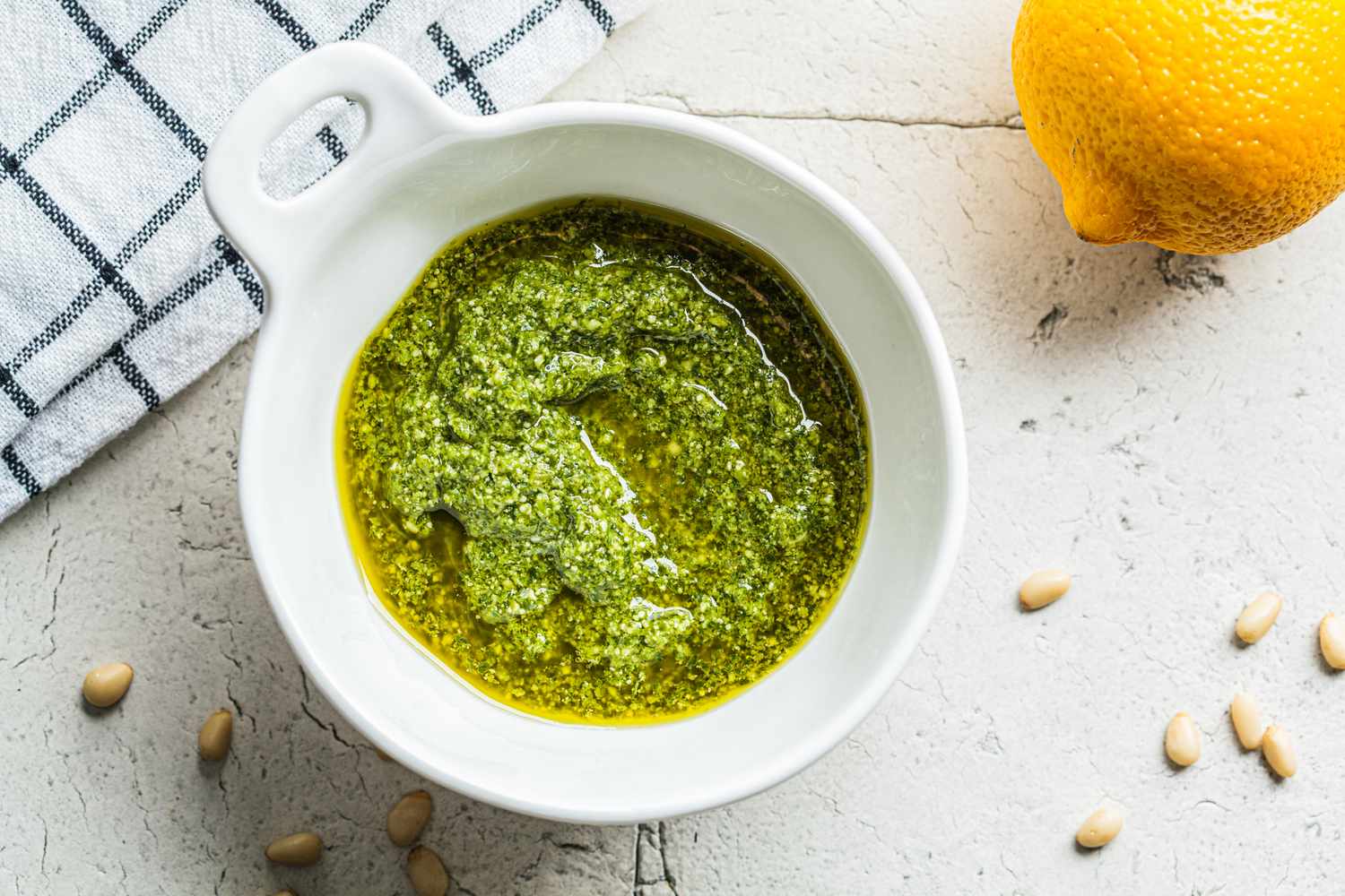 Bowl of pesto next to a lemon and scattered pine nuts on a countertop