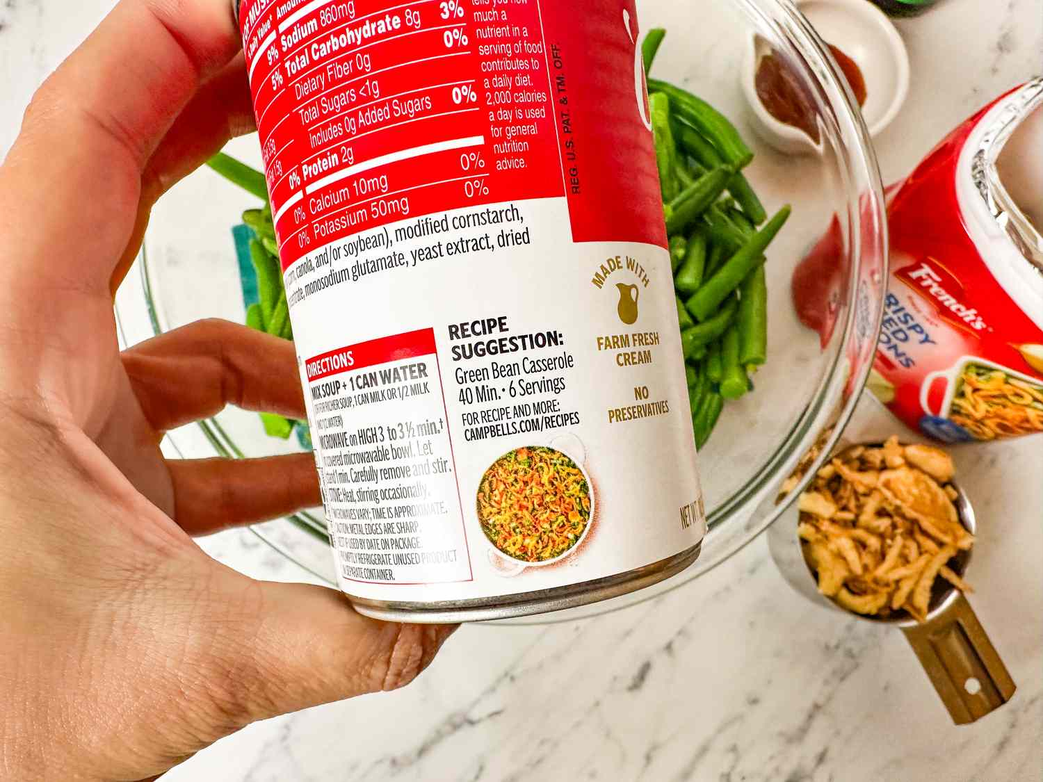 Hand holding a can of Campbells soup with green beans and other ingredients visible on a marble countertop