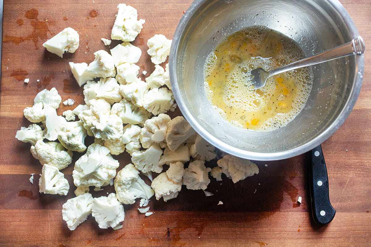 A metal bowl with egg and seasonings mixed with a fork and set next to cauliflower florets to make Air Fryer Cauliflower with Buffalo Sauce.