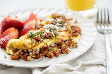 Horizontal image of the best corned beef omelet. The folded omelet is covered in browned corned beef. Grated cheese and parsley are on the top of the omelet. A white cotton napkin is partially under the plate. A fork rests on the napkin which is to the right of the plate. A partial view of a glass of orange juice is above the plate. Halved strawberries are to the left of the omelet on the plate.