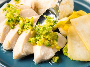 A plate of sliced chicken topped with a scallion sauce, served with rice and pickled vegetables
