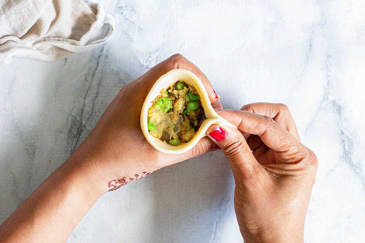 Woman pinching together a cone of dough filled with vegetables in her hands ot make Indian Samosas.