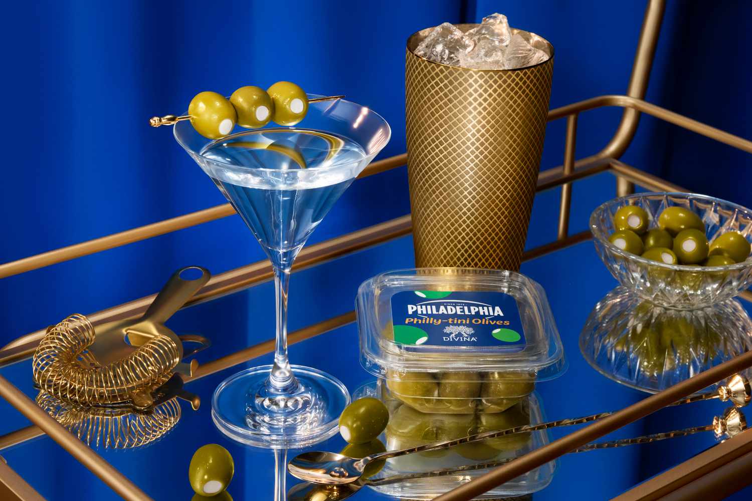 A martini with olives and a container of Philadelphia brand olives on a gold bar cart surface