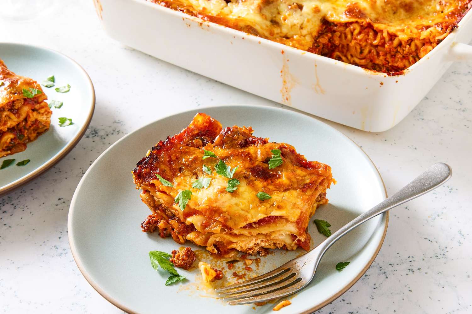 A portion of lasagna served on a plate with a fork garnished with herbs next to a baking dish containing the remaining lasagna