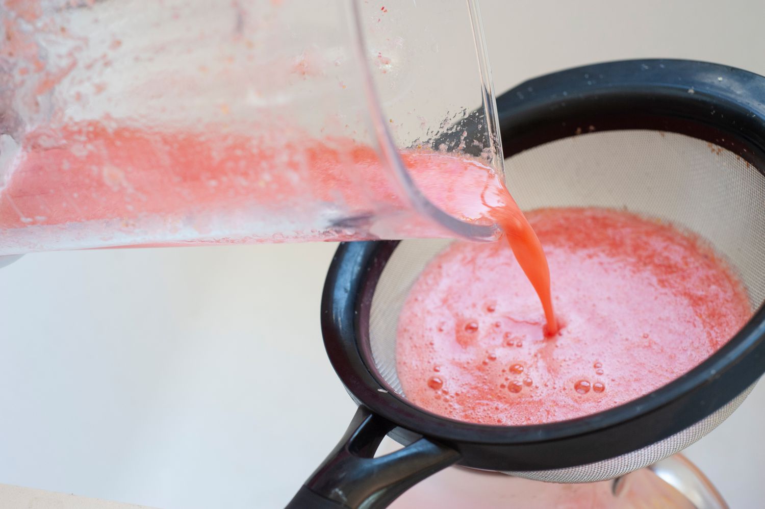 Pouring blender strawberry lemonade through a fine-mesh sieve.