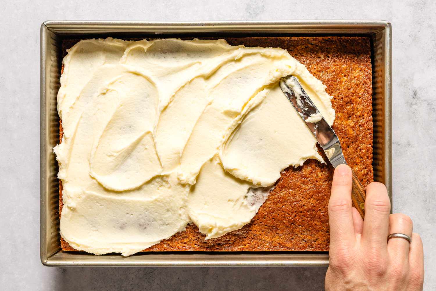 Spreading frosting on a rectangular carrot cake in a pan from Easy Carrot Cake recipe, hand holding an offset spatula
