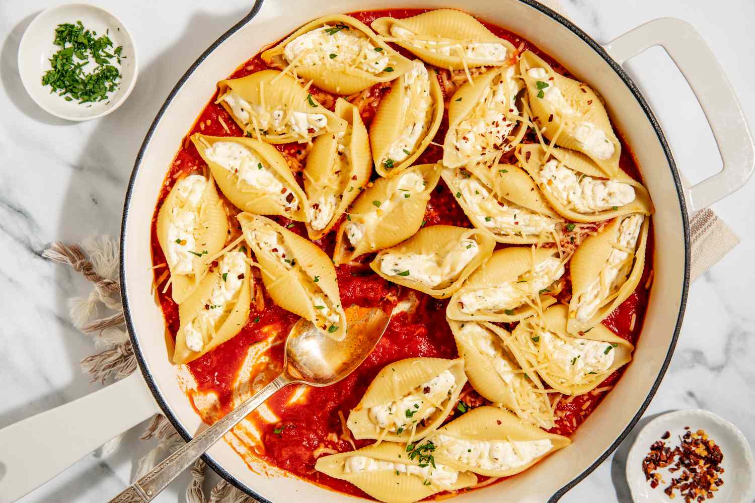Stovetop Stuffed Shells in a large pan with a large serving spoon next to small dish with crushed red pepper and small dish with chopped parsley