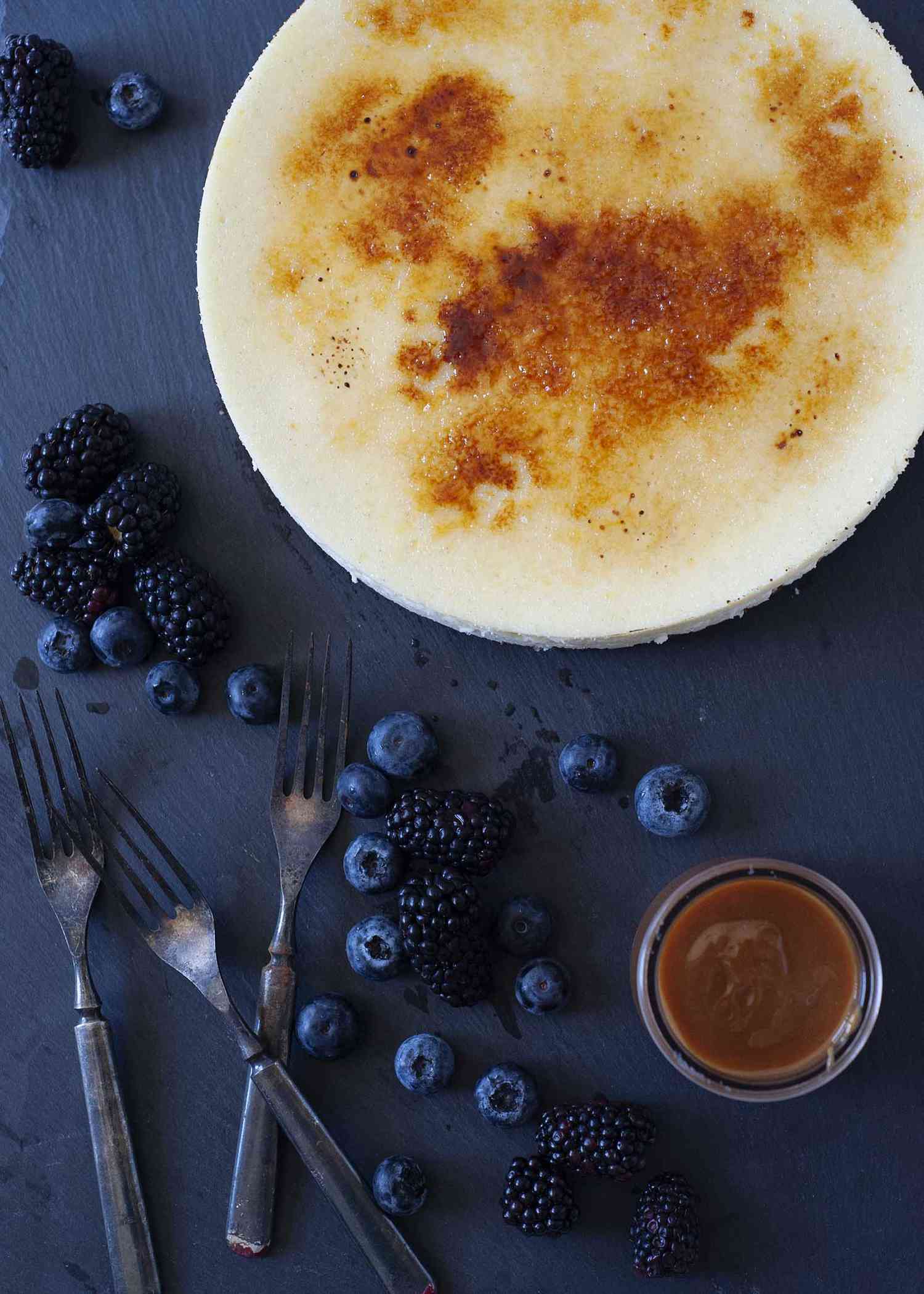 Vanilla Bean Cheesecake with brittle sugar topping on a slate board wtih fruit and caramel nearby.