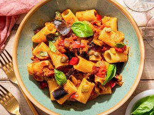 caponata pasta in a bowl (close up) next to a glass of wine, utensils on the table, and a small bowl of basil, all at a rustic table setting 