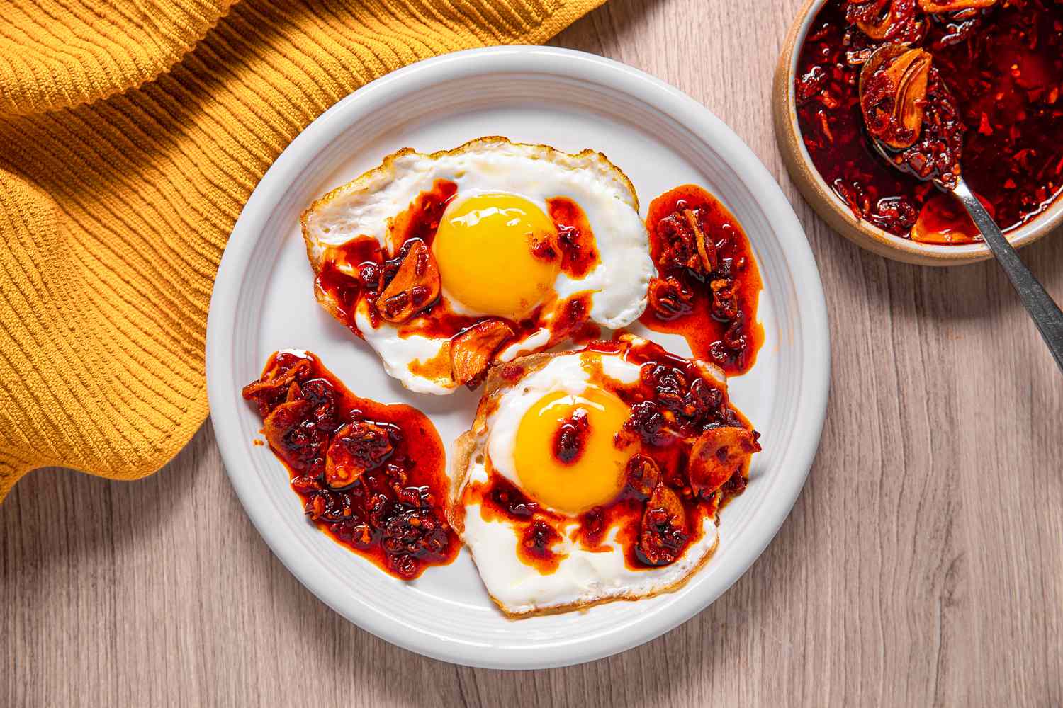 Sunny Side Eggs Topped With Homemade Chili Crisp, and in the Surroundings, a Mustard Colored Kitchen Towel and a Bowl With More Chili Crisp