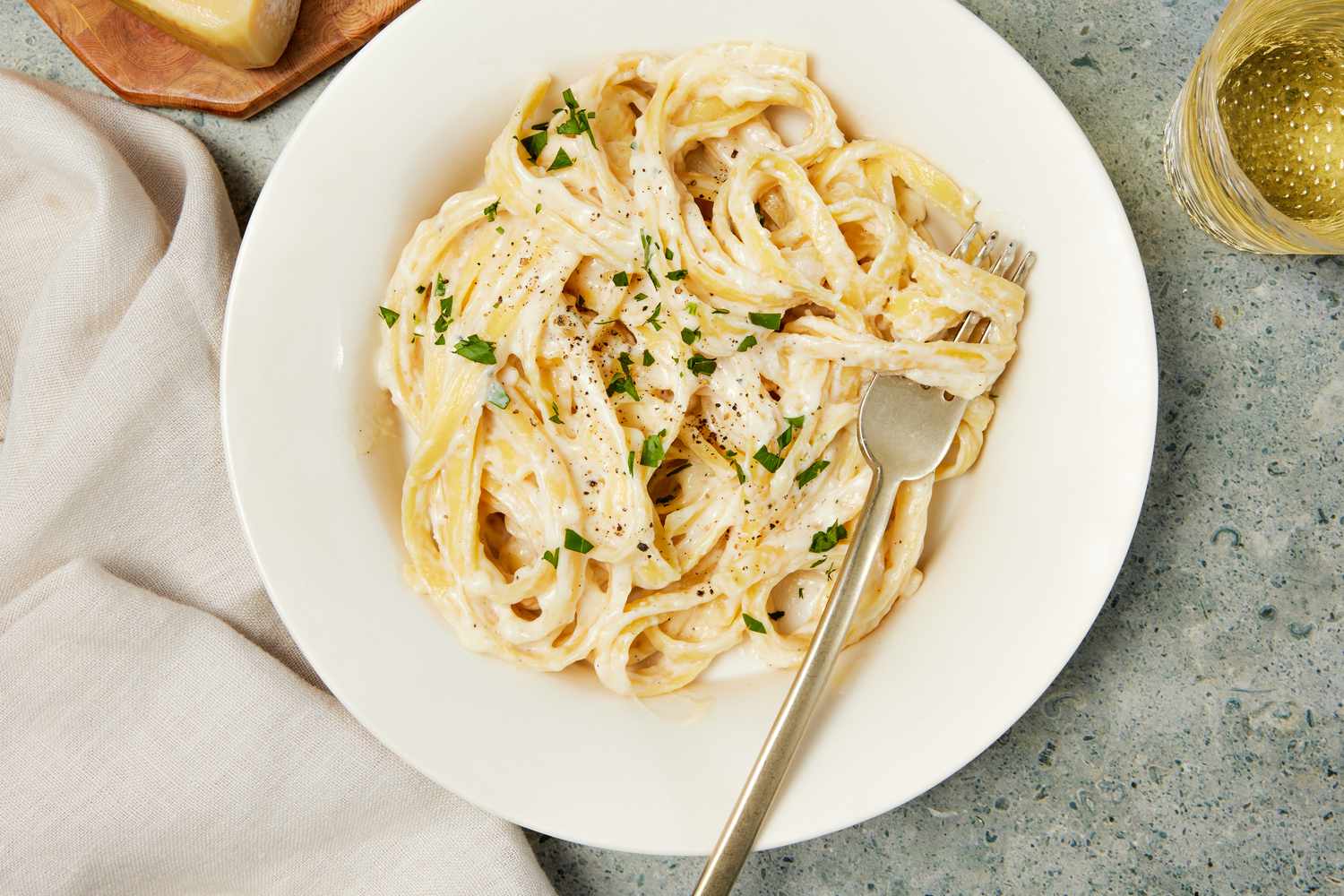 Copycat Olive Garden alfredo in a bowl with a fork, and in the surroundings, a white table napkin, a block of parmesan on a wooden board, and a glass with a drink
