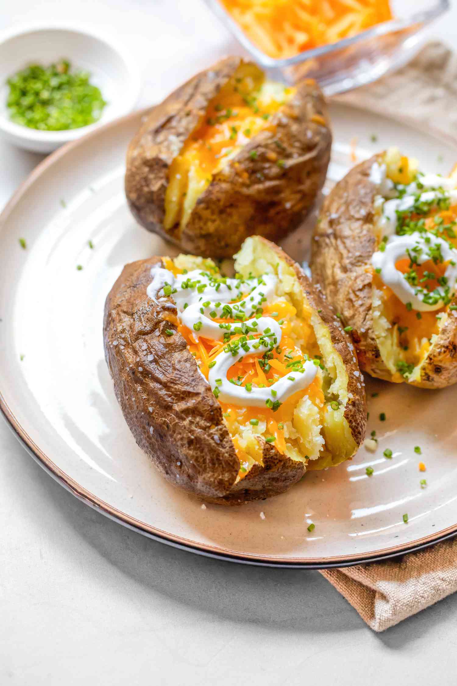 Microwave Baked Potatoes Topped with Sour Cream, Cheddar Cheese, and Chives on a Plate, and in the Background, a Small Plate with More Cheese and a Saucer with More Chives