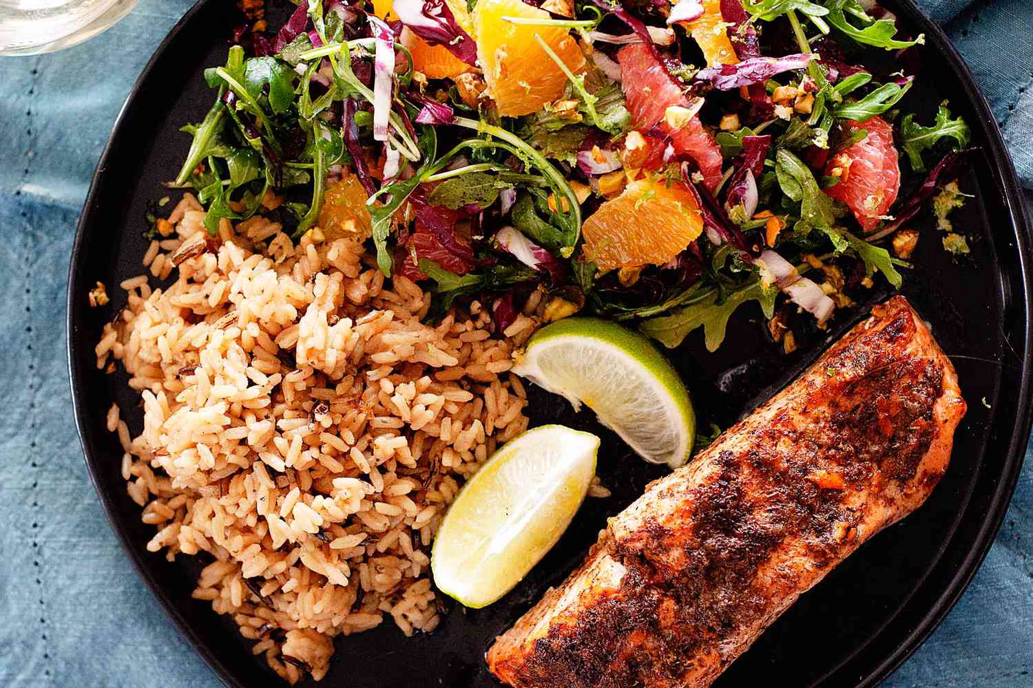 A blackened fillet of weeknight jerk salmon is on the lower right of a black plate. Lime wedges, red rice and a mixed green salad with citrus mixed in is to the left of the salmon. The plate rests on a blue linen.