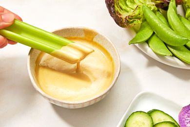 A stick of celery being dipped into a bowl of creamy mustard maple sauce, accompanied by fresh vegetables including snap peas and sliced cucumber