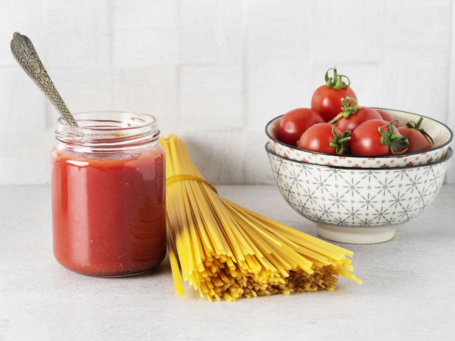 Dried spaghetti on a counter with a jar of tomato sauce and a bowl with fresh toamtoes