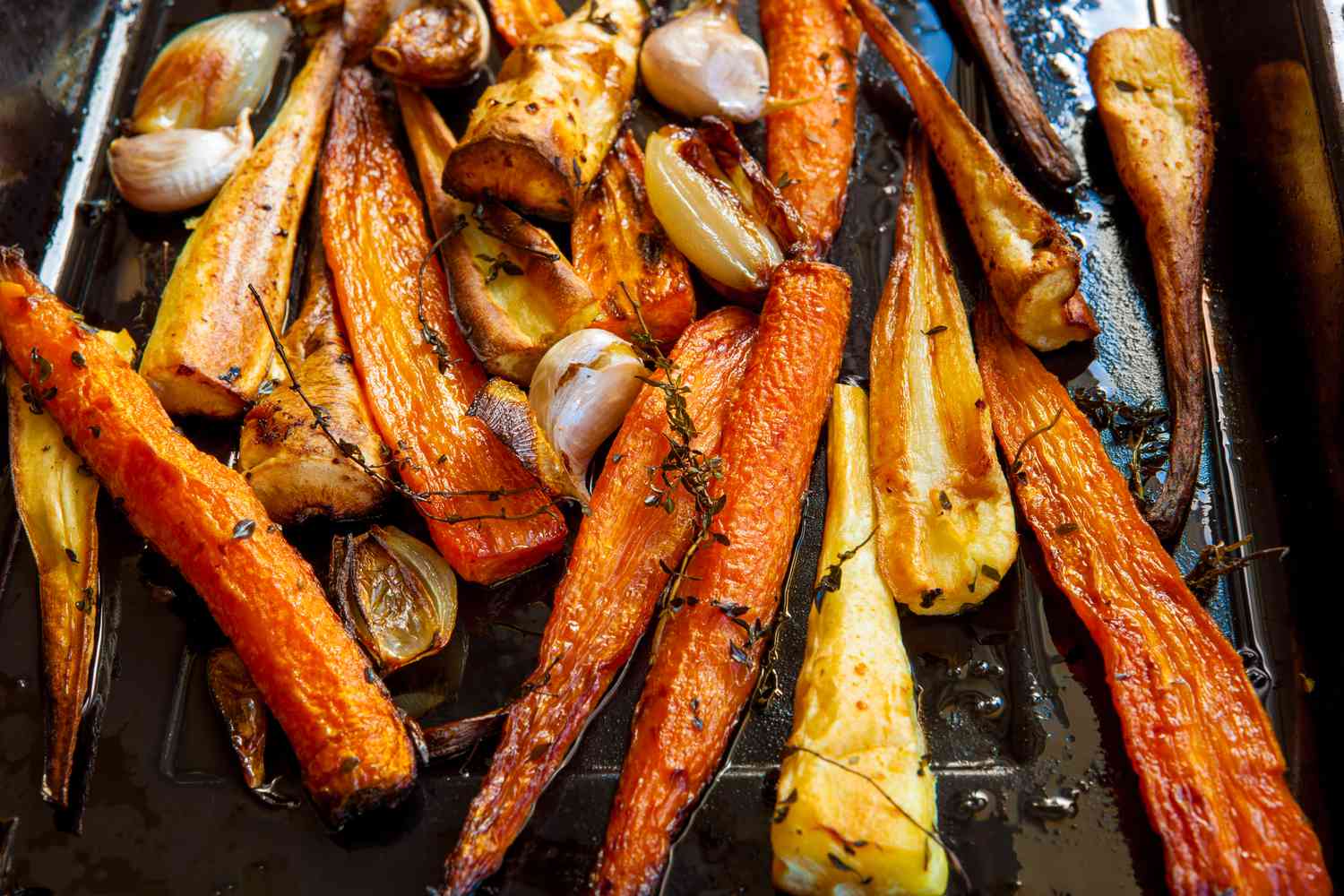 Overhead view of carrots and parsnips roasting in a pan, with thyme, shallots, and garlic.