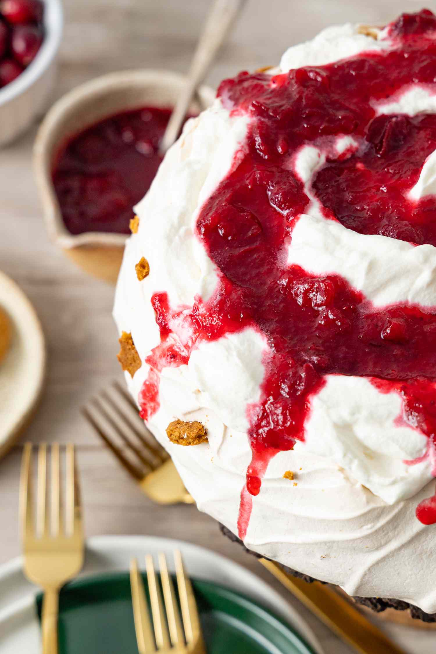 Gingersnap Pavlova with Cranberries, and in the Background, Forks on the Counter and on a Stack of Plates, a Bowl of More Sauce, and a Bowl of Cranberries