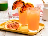 Two Salt Rimmed Glasses of Grilled Grapefruit Paloma With Two Grilled Grapefruit Slices on the Glass Rim Next to More Slices of Grilled Grapefruit on a Small Tray, and in the Surroundings, a Bottle of Club Soda and a Cream Pitcher