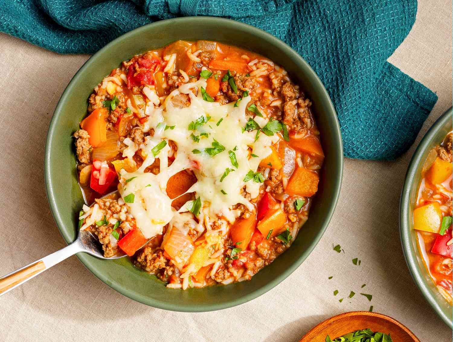 Bowl of stuffed pepper soup topped with cheese and some of the soup in a spoon being taken from the bowl, and in the surroundings, a table setting with a table napkin, another bowl of soup, and a small bowl of herbs