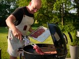 Person grilling meat using tongs on a charcoal grill in a garden setting on a sunny day, wearing an apron and holding a tray