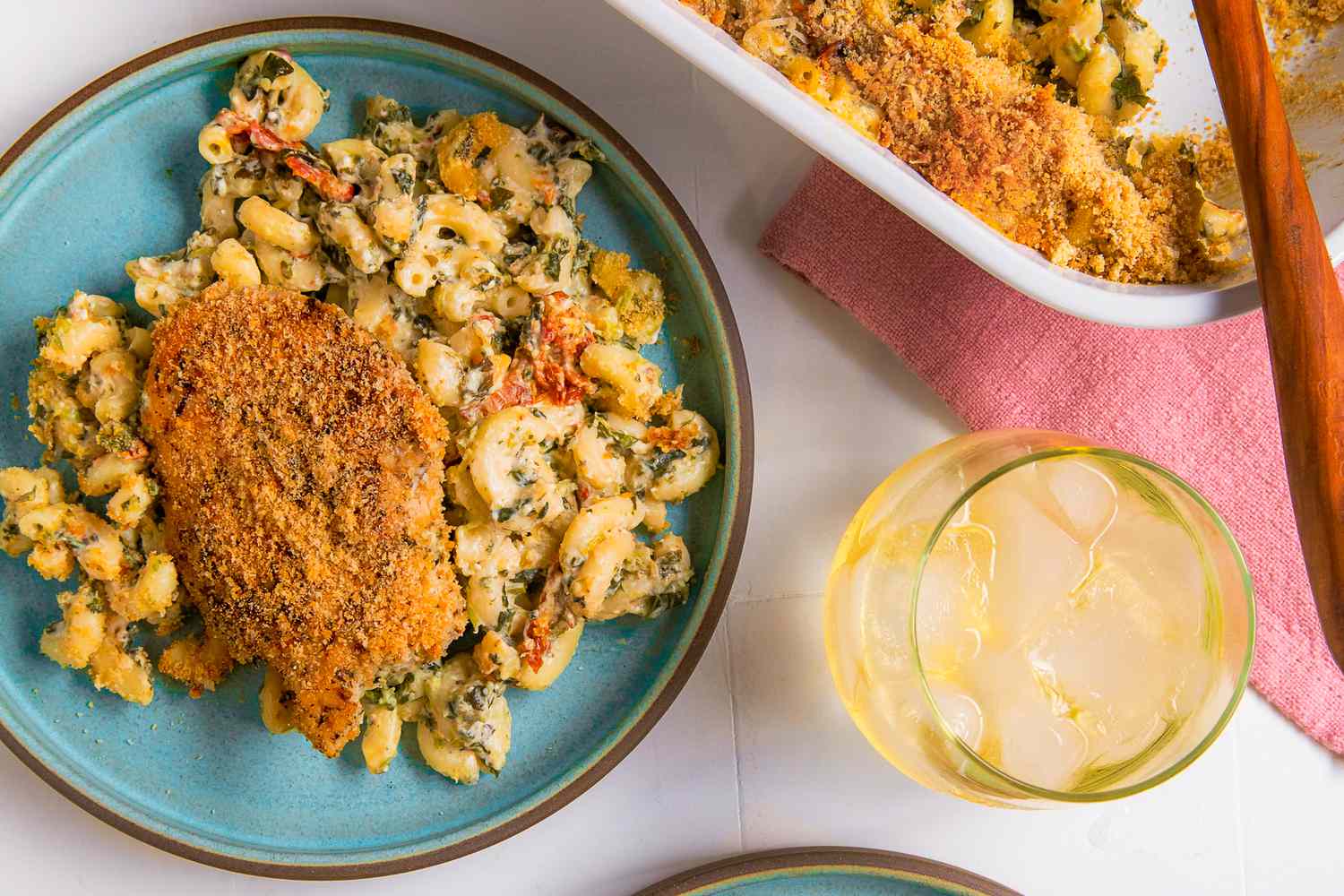 Overhead view of chicken and pasta casserole serving on a blue plate next to a dish of casserole, table napkin and drinking glass with ice and beverage