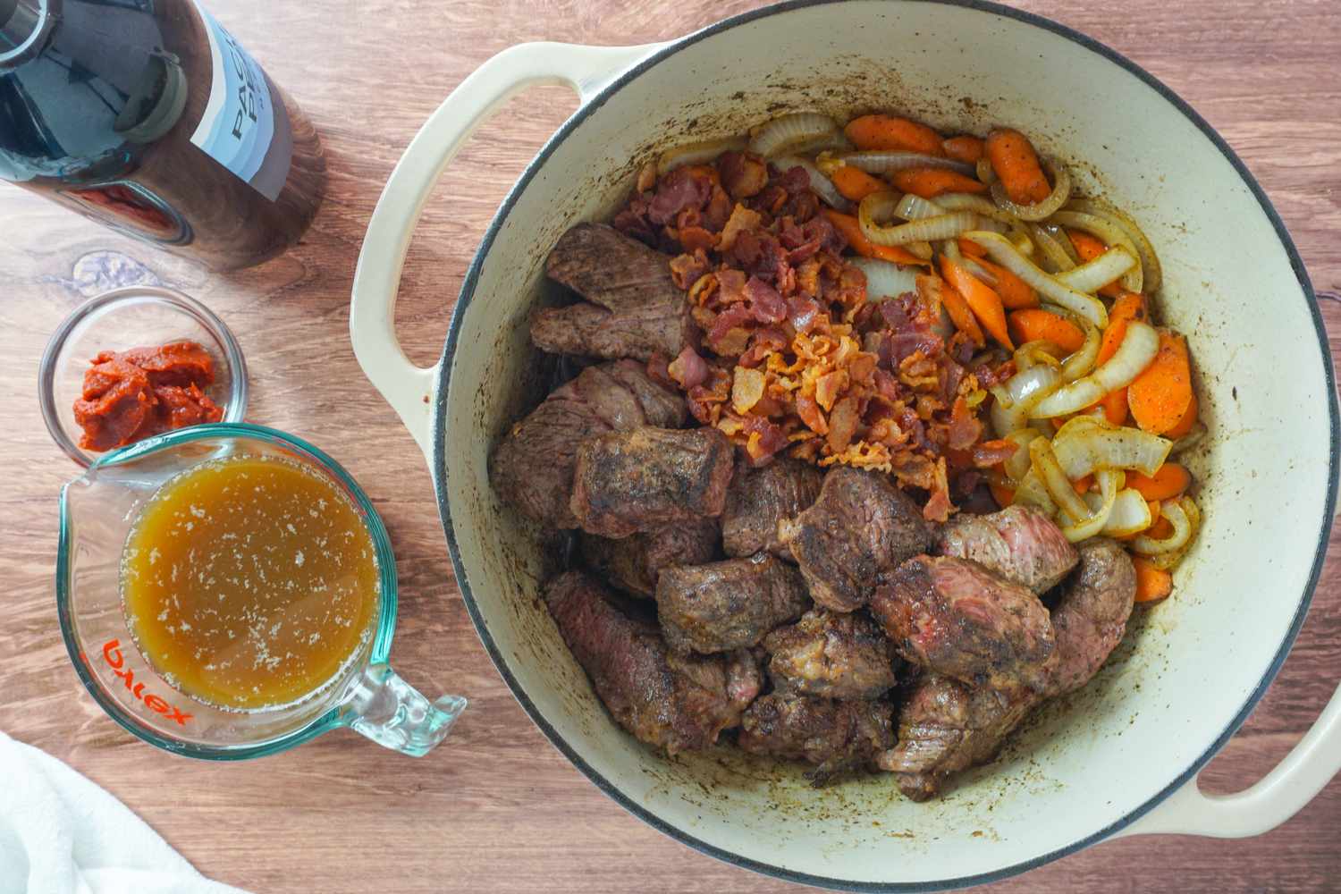 A pot with beef stew ingredients alongside stock in a measuring cup and tomato paste in a bowl on a wooden surface