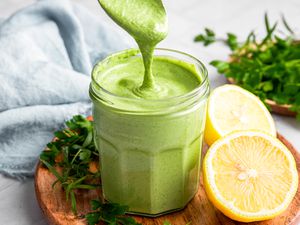 Green Goddess Dressing Dripping from a Spoon into a Jar with More Dressing. Jar is on a Wooden Plate with Herbs and Halved Lemons