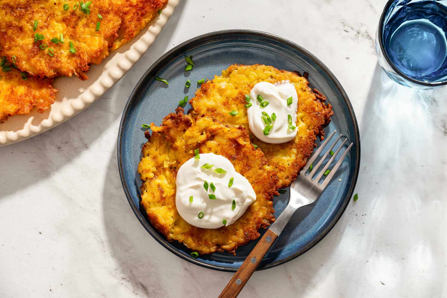 Plate with two crispy latkes topped with sour cream and chives accompanied by a fork on the side