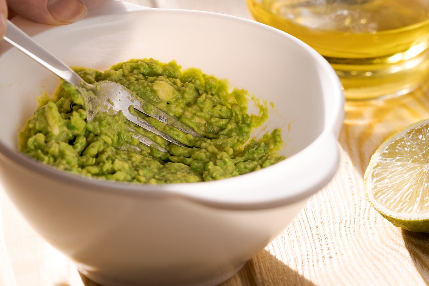 White bowl with fork preparing homemade guacamole from organic produce with a lime and bottle of oil to the side