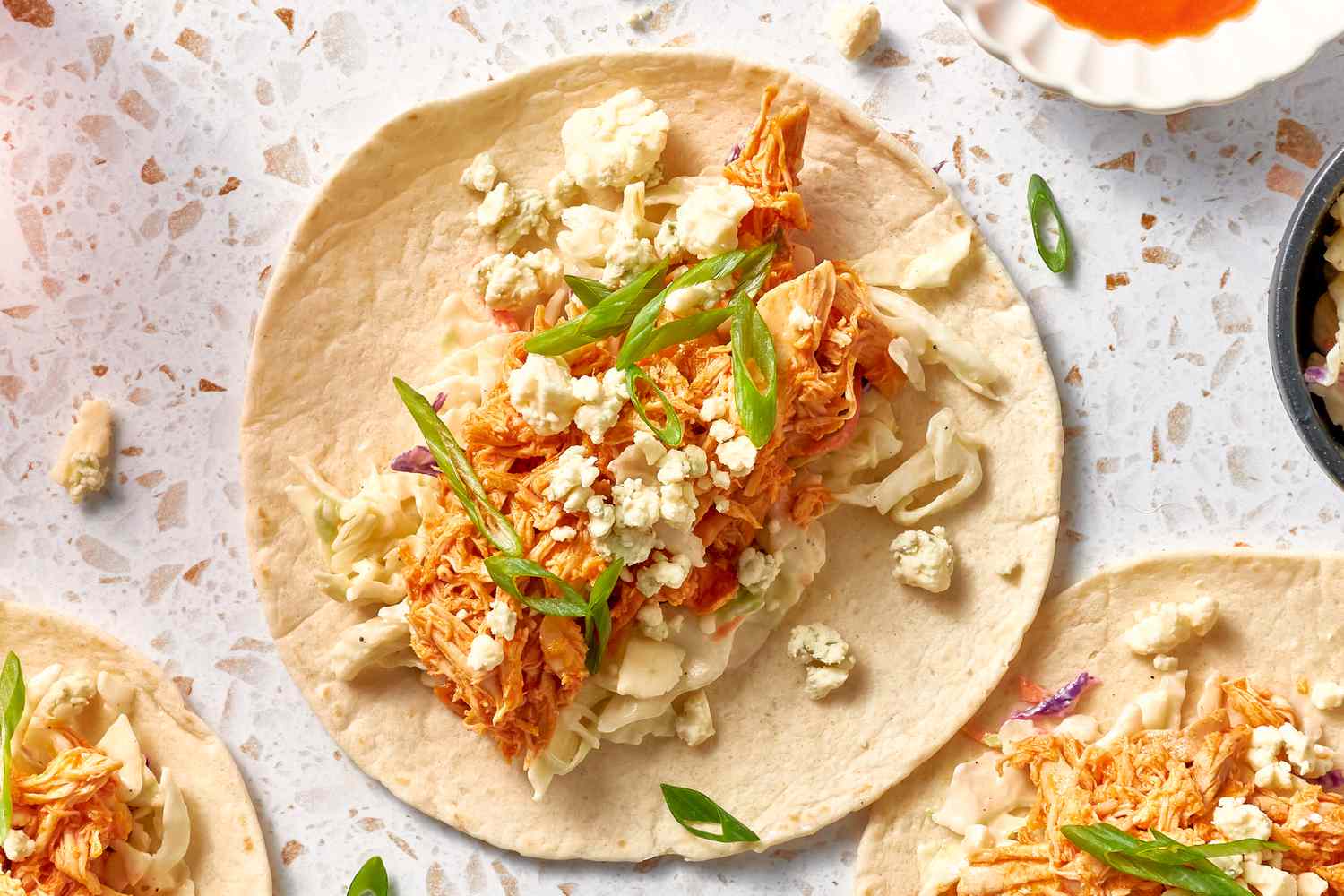 Buffalo Chicken Tacos Topped With Blue Cheese Crumbles and Sliced Green Onion Next to More Buffalo Chicken Tacos, a Small Bowl Witch Chicken and a Small Bowl With More Buffalo Sauce