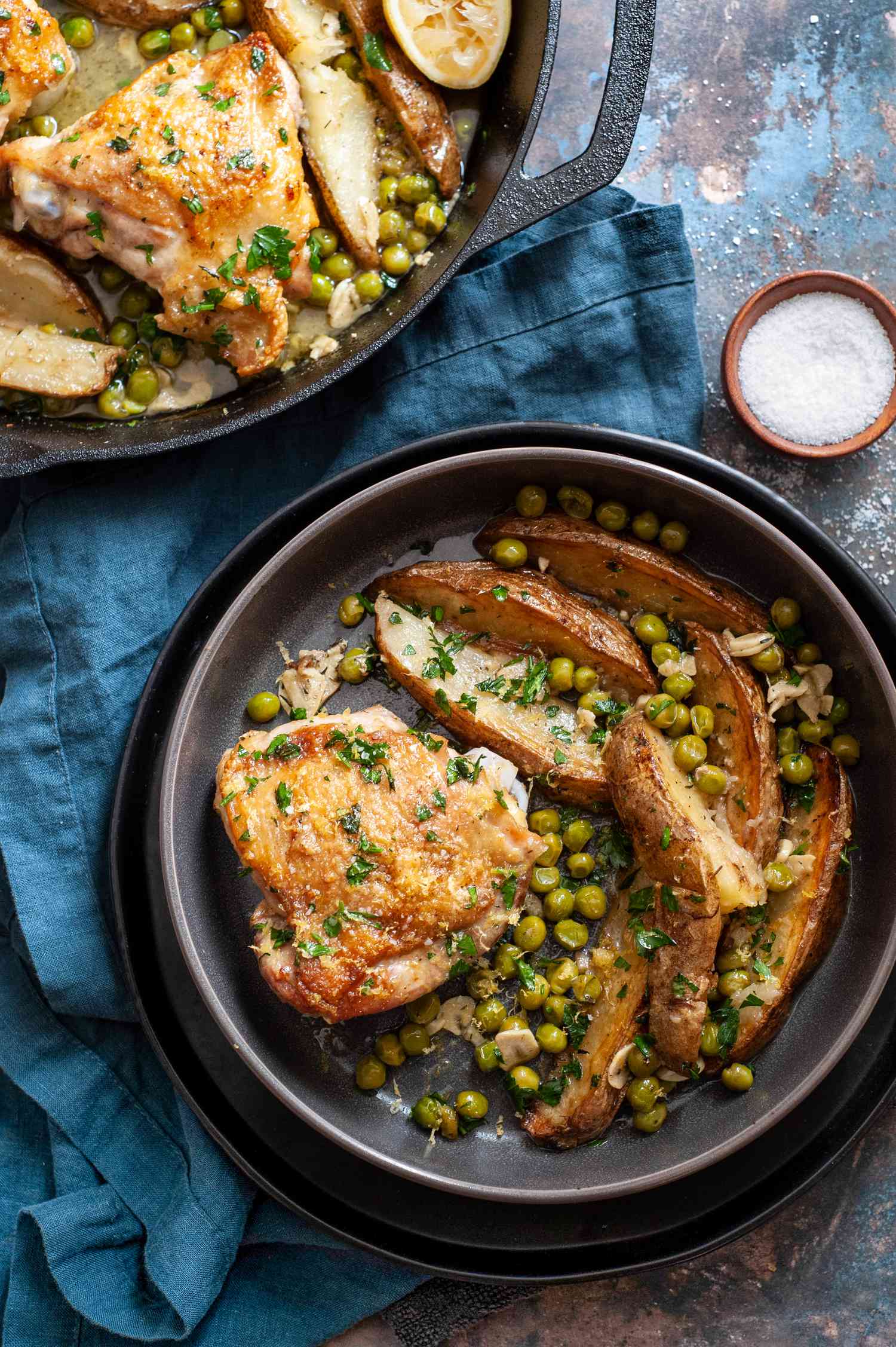 Chicken Vesuvio in a Bowl on a Plate Next to a Cast Iron Skillet with More Chicken Vesuvio and a Small Bowl of Salt, All Sitting on a Table Napkin