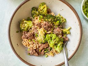 Overhead view of a brown rustic style bowl of ground beef, broccoli, noodles and a fork next to a small bowl of chopped green onions on a light blue background