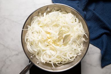 Sliced Onions, Garlic, and Bay Leaves in a Pan on a Portable Stove and Next to a Blue Kitchen Towel for French Onion Meatballs
