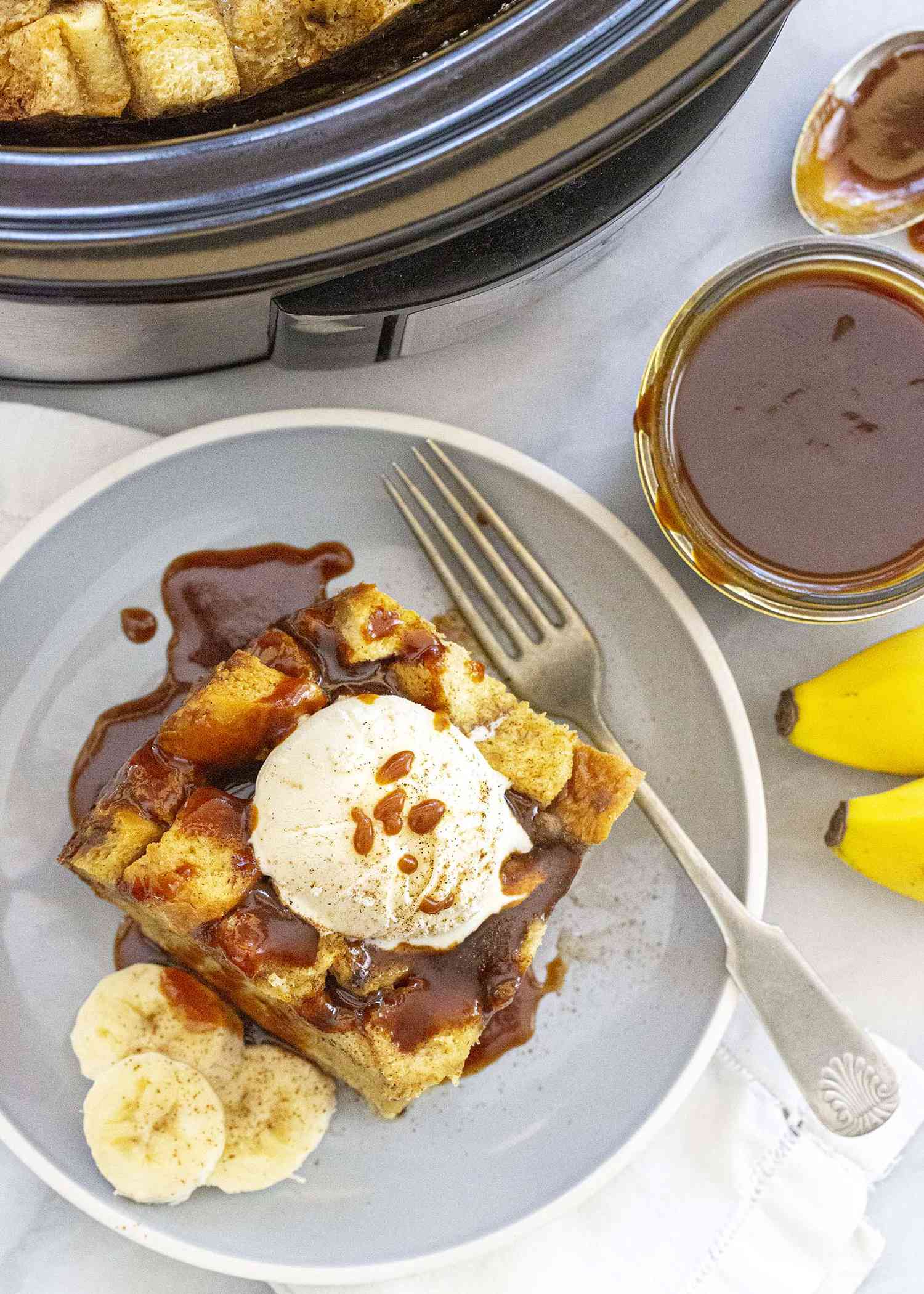 Slice of easy to make slow cooker banana bread pudding topped with a scoop of ice cream and drizzeld with caramel sauce on a gray plate with a fork and sliced bananas. The slow cooker and a jar of caramel sauce is off to the side.