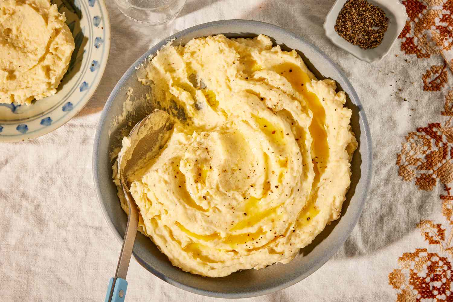 Overhead view of a gray bowl of almond milk mashed potatoes with a spoon and topped with butter on top of a fall themed embroidered cloth