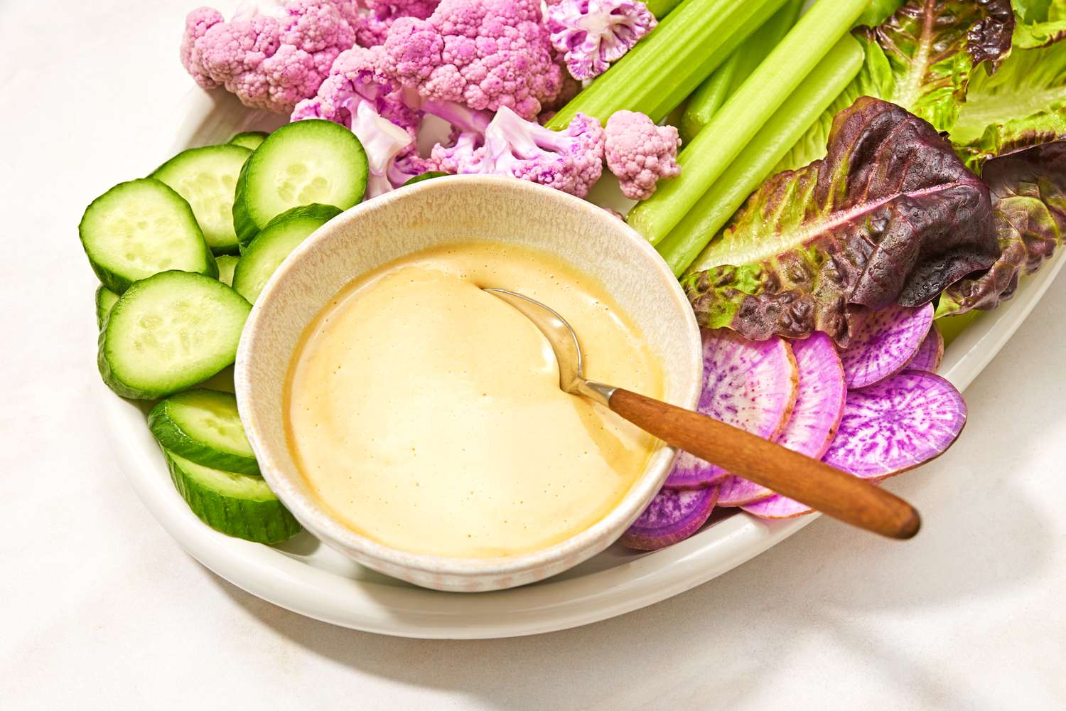 A bowl of creamy mustard maple sauce with a spoon, surrounded by fresh vegetables on a plate including cucumbers, radishes, celery, cauliflower, and lettuce