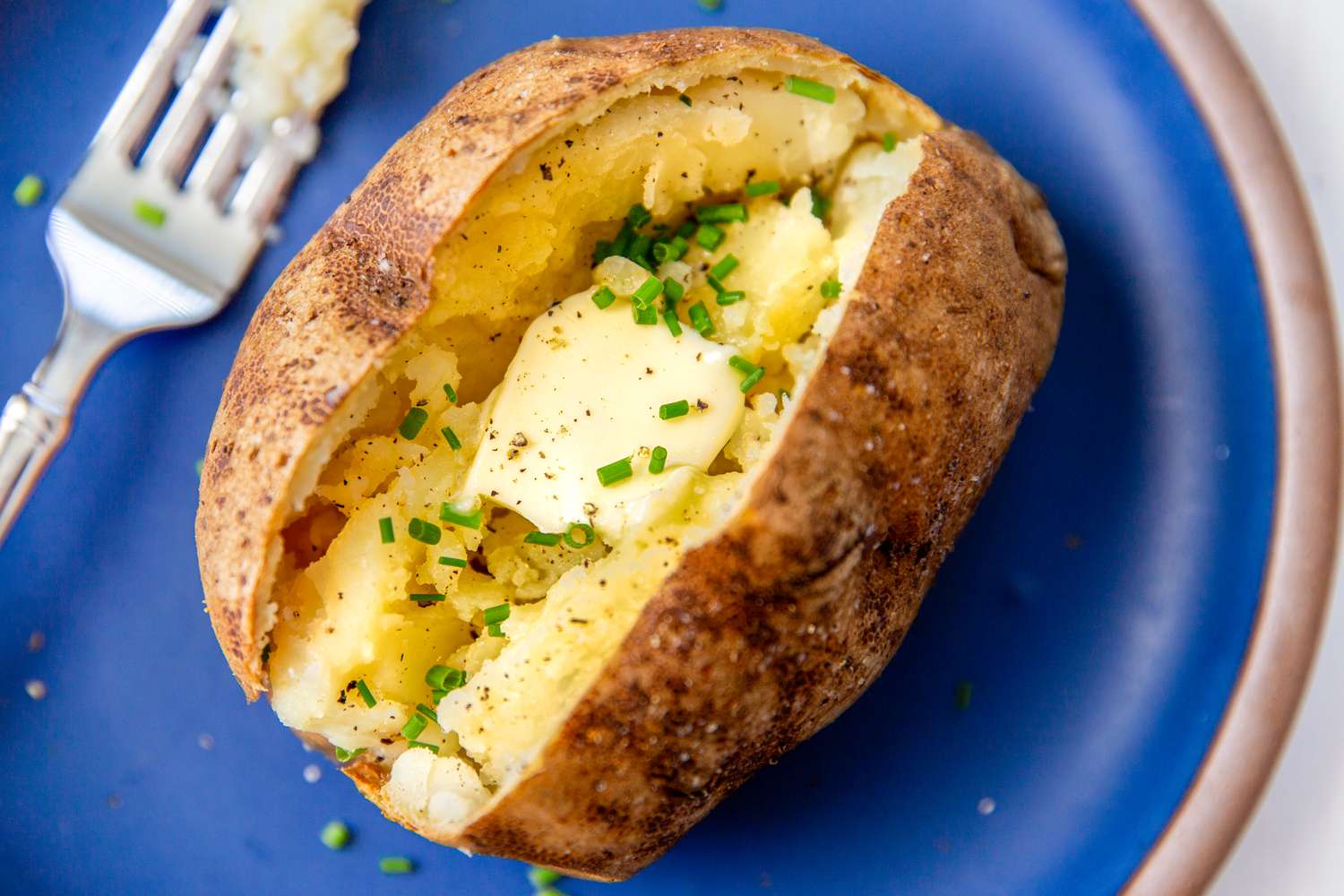 baked jacket potato cut open length wise and filled with butter and chives, on a blue pate with a fork on the left side
