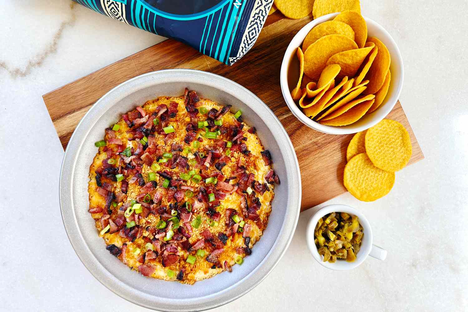 A bowl of cheesy dip garnished with green onions and bacon accompanied by a bowl of tortilla chips and a small dish of jalapeos