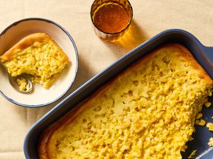 Overhead view of a dark blue baking dish of corn soufflé with a serving removed and a white bowl and spoon with a serving next to a drinking glass
