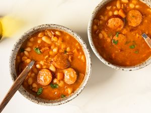Two bowls of bean and sausage stew on a white surface wooden spoons in each bowl