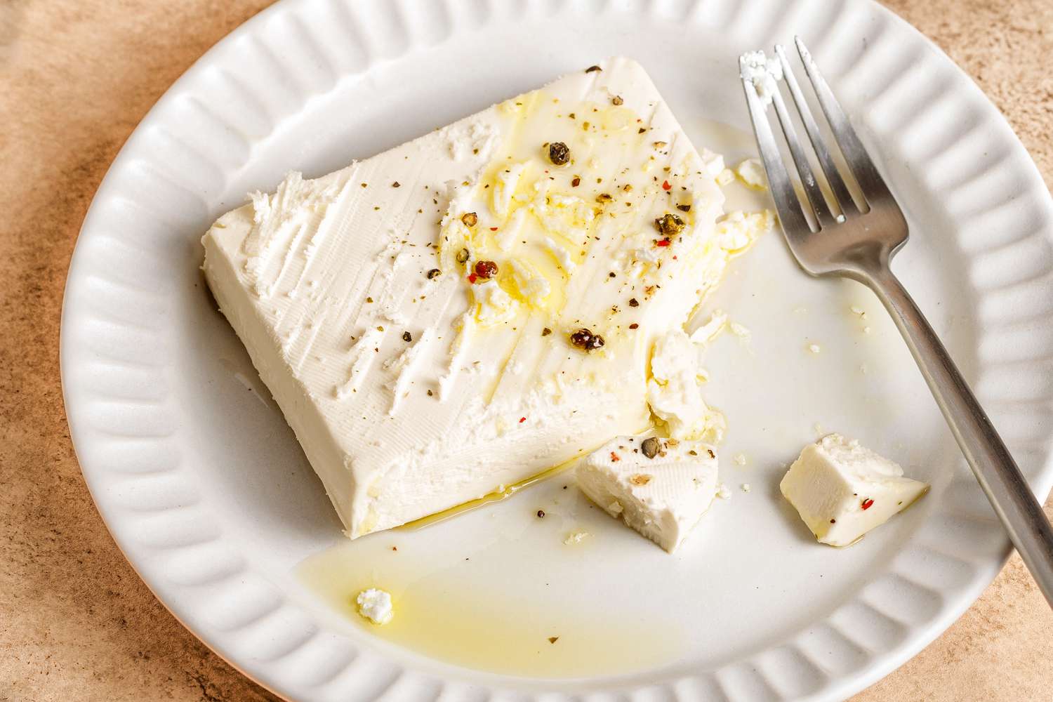 A block of feta cheese garnished with seasoning and olive oil on a white plate with a fork