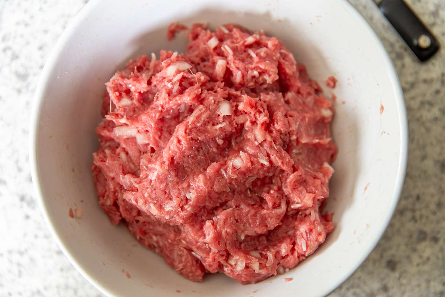 Overhead view of a white bowl of raw ground beef on a granite countertop