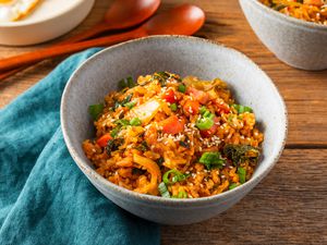 Bowl of kimchi and spam fried rice at a table setting with another serving in a bowl, a table linen, two spoons, and a plate with a fried egg