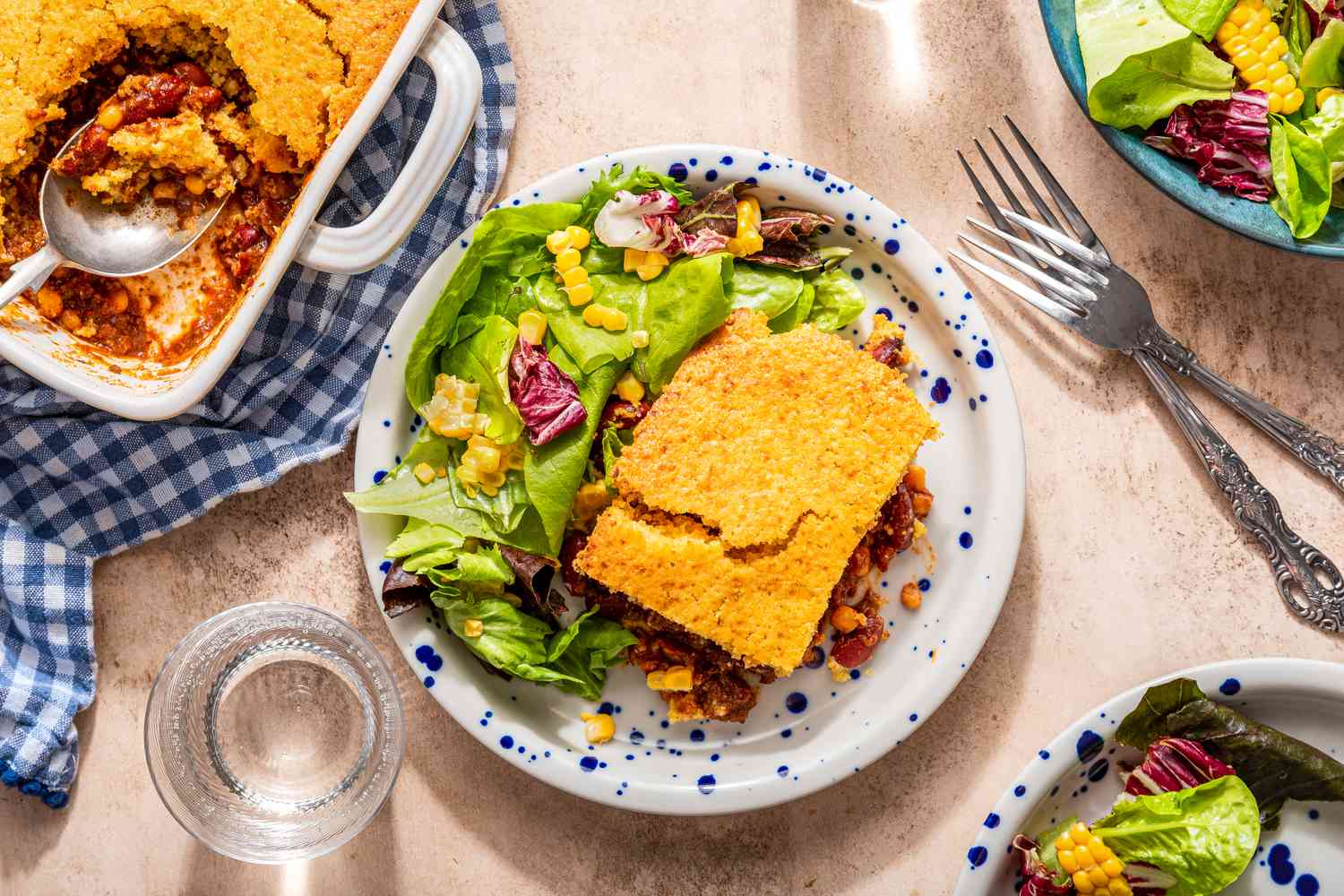 overhead shot of upside down chili pie on a plate with a side salad at a table setting with a casserole dish with more, a bowl with more salad, and a plate with another serving of chili pie