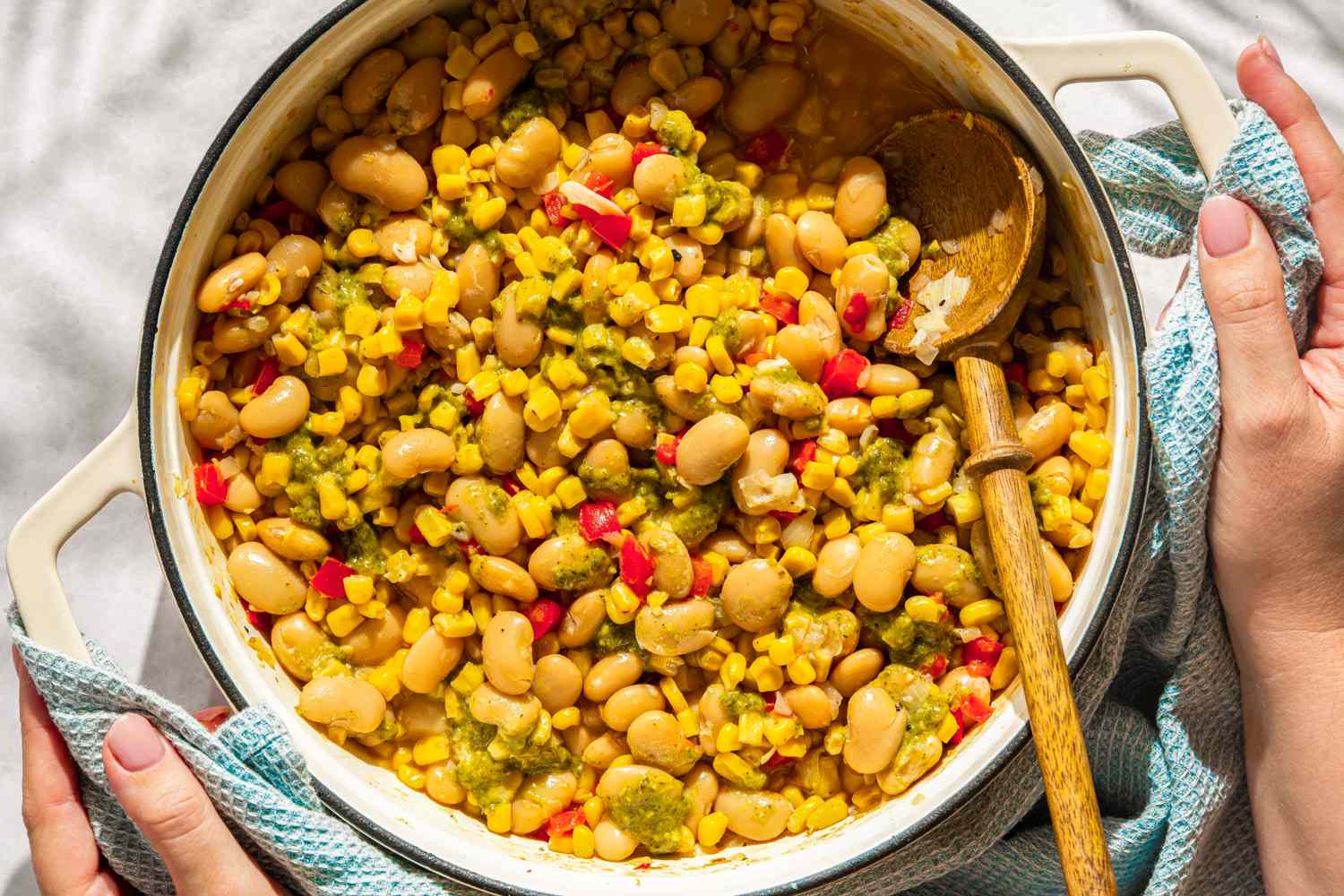 Pot of mixed vegetables including corn, beans, and peppers, with a wooden spoon and hands holding the pot