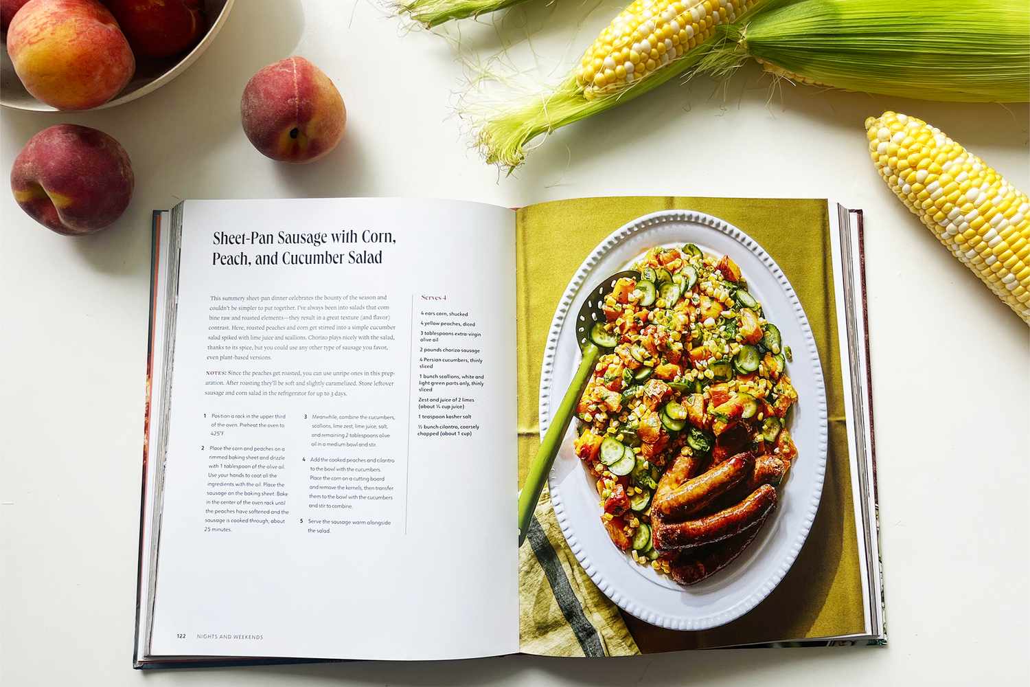 Open cookbook showing a recipe for sheet pan sausage with a salad surrounded by peaches and corn on the cob